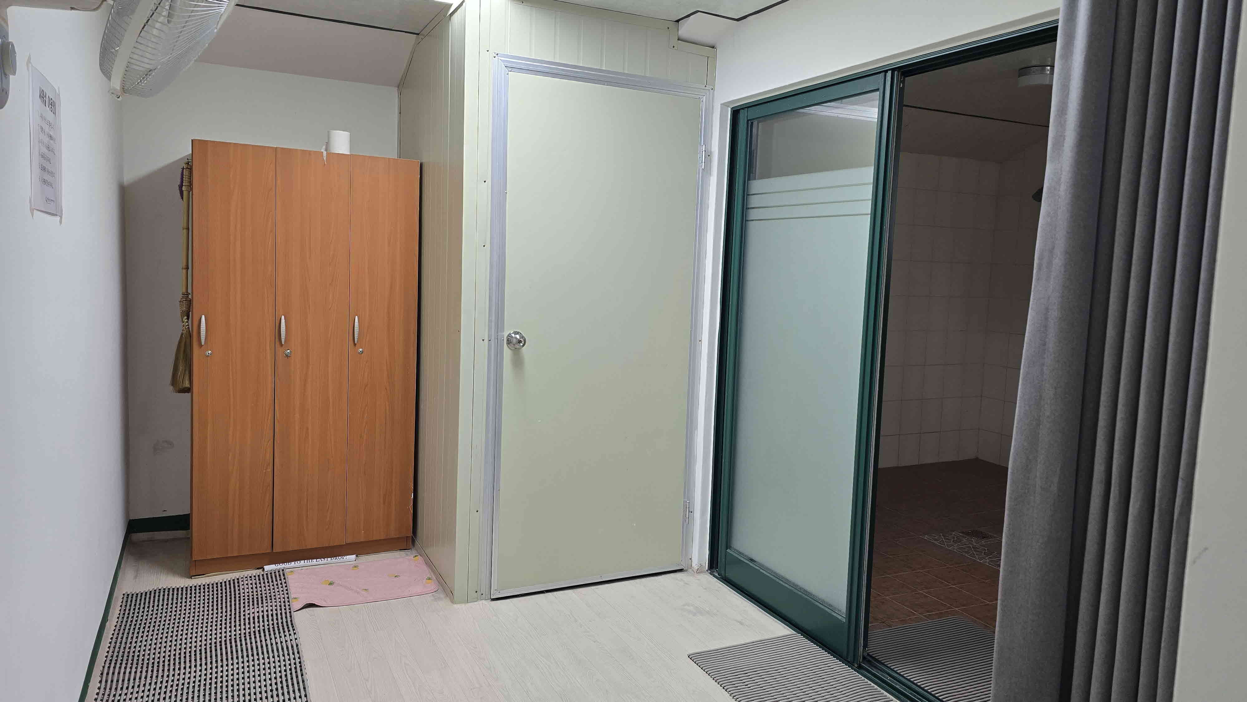 샤워실 앞 탈의실 내부에서 촬영한 사진이며, 좌측에는 선풍기와 목재 캐비넷이 비치되어 있고, 우측에는 샤워실로 향하는 문이 위치해 있습니다.