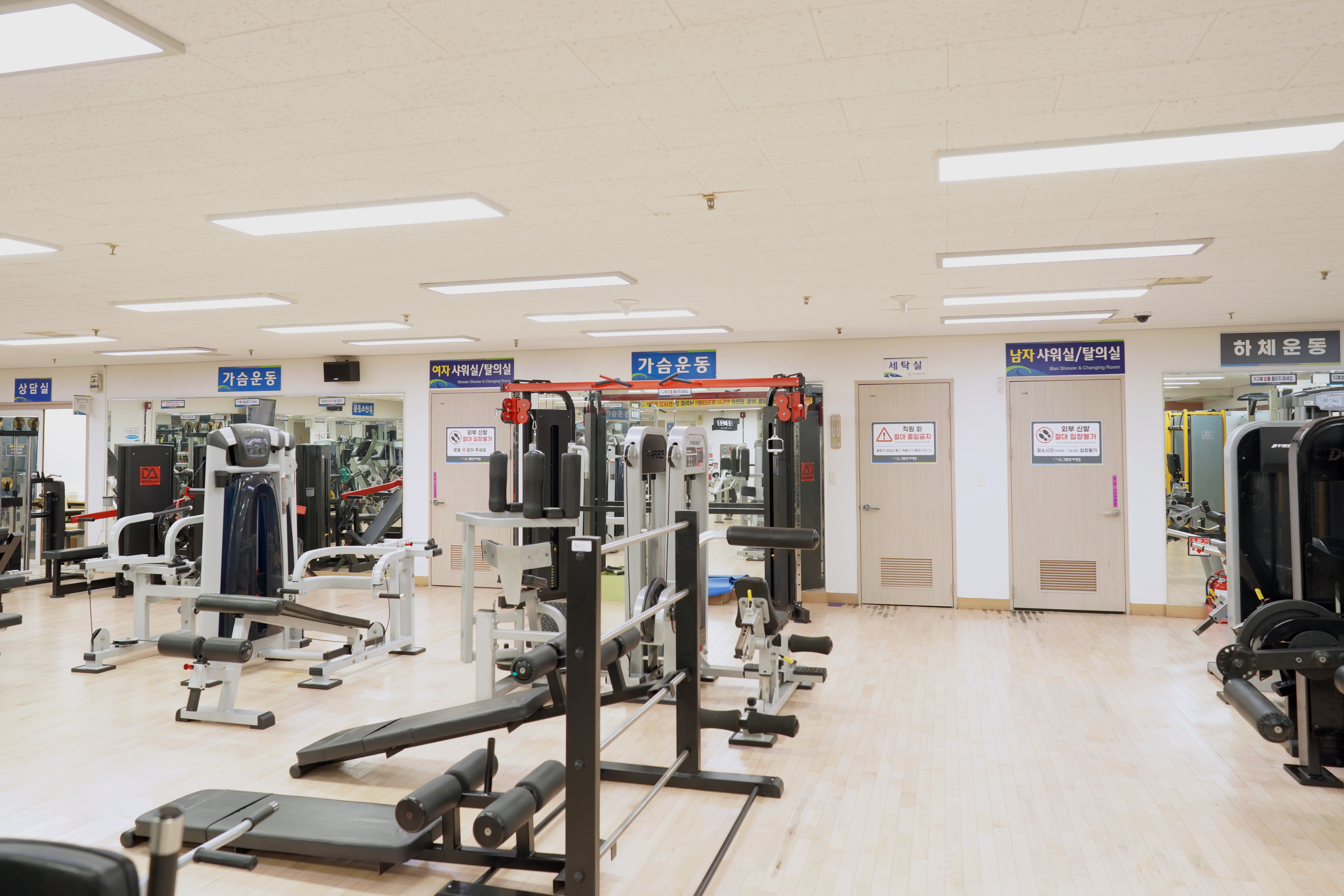 창원실내수영장 지하 웨이트실을 정면에서 촬영하여 운동기구 배치와 가슴 운동, 하체 운동 등의 섹션 표지판을 포함한 시설 구성을 담은 사진입니다.