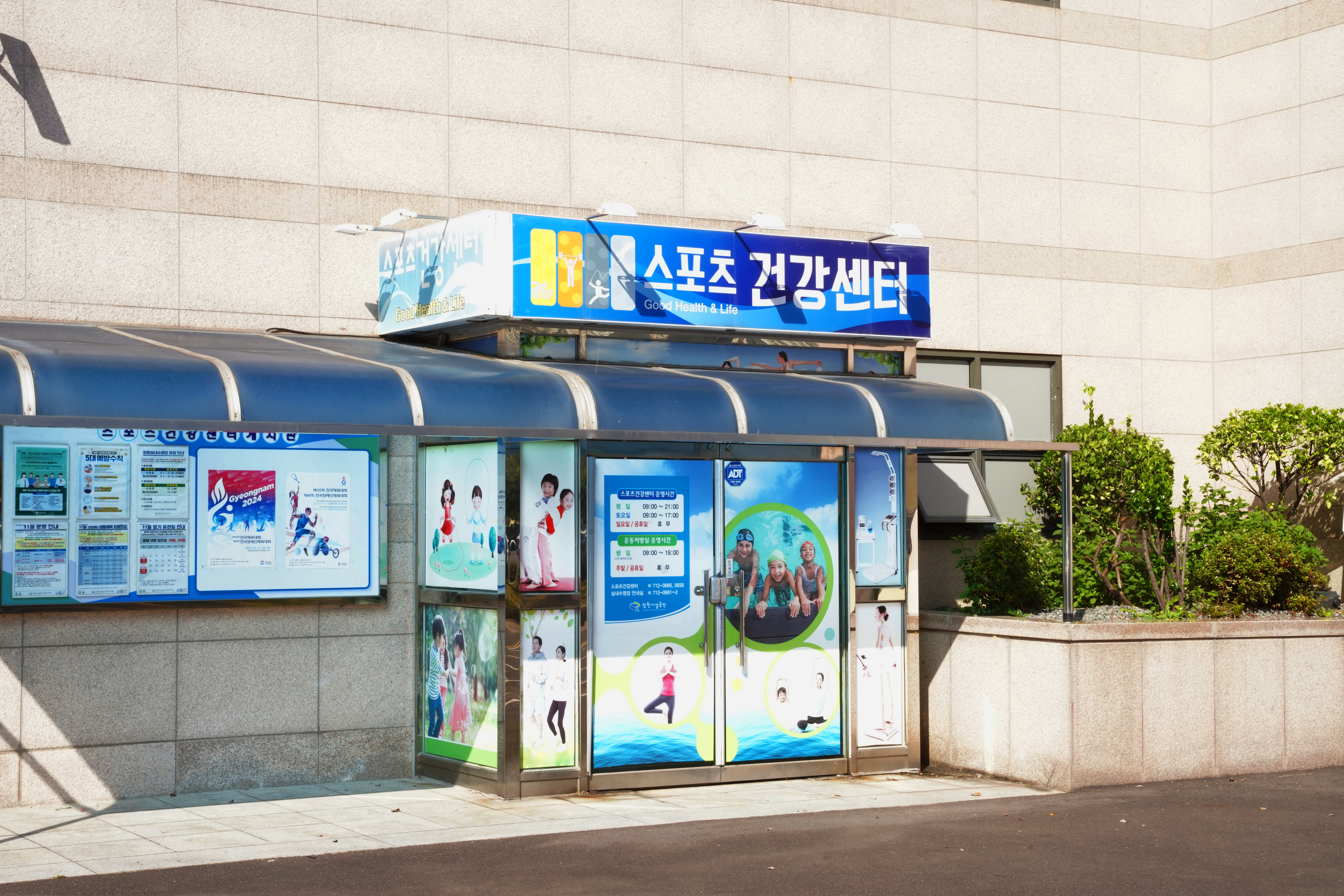 창원실내수영장 스포츠 건강센터 입구를 측면에서 촬영하여 간판, 게시판, 외부 디자인, 주변 조경을 담은 사진입니다.