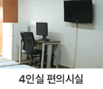 4인실 편의시설 : 창원축구센터 숙소동 4인실의 편의시설을 찍은 사진입니다. 밝은 색 벽지 앞에 철제 프레임으로 된 책상과 컴퓨터, 의자가 중앙에 배치되어 있고, 왼쪽으로는 냉장고와 우드톤의 캐비닛이 배치되어 있습니다. 우측 벽면에는 벽걸이 TV가 걸려있습니다.