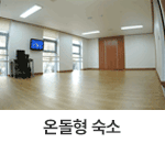 온돌형 숙소 : 창원축구센터 온돌형 숙소의 사진입니다. 창이 3개나 있어 채광이 좋고, 넓은 공간이 특징입니다. 안쪽에는 캐비닛이 위치하고 있어 침구류, 의류를 보관할 수 있고 창 사이에 책상과 컴퓨터, TV가 위치하고 있어 작업공간으로 활용할 수 있습니다. 밝은 하얀색 벽지와 우드 톤의 바닥이 따뜻한 느낌을 주는 공간입니다.