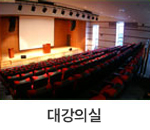 대강의실 : 대세미나실의 내부를 뒤에서 바라본 모습으로 강의실 앞쪽에는 대형 스크린과 연단이 있으며, 연단 옆에는 발표자가 사용할 수 있는 책상이 배치되어 있습니다. 좌석은 계단식으로 배치된 빨간색 의자로 구성되어 있고, 여러 줄로 줄지어 있습니다.