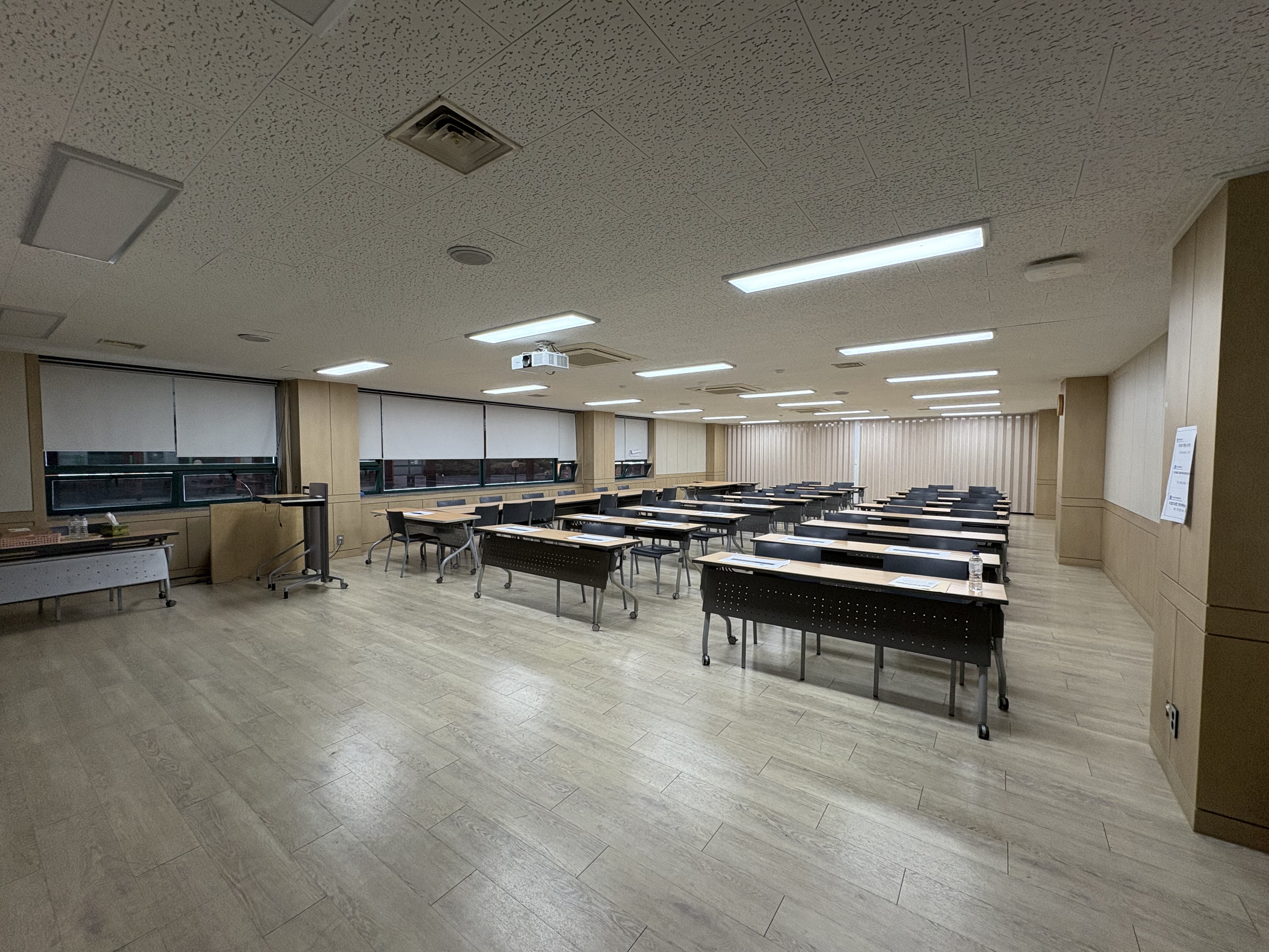 중강당 바깥쪽 가장자리에서 안쪽을 바라보며 촬영한 사진입니다. 2인용 책상과 의자들이 3열로 나란히 놓여있는 전면이 보입니다.