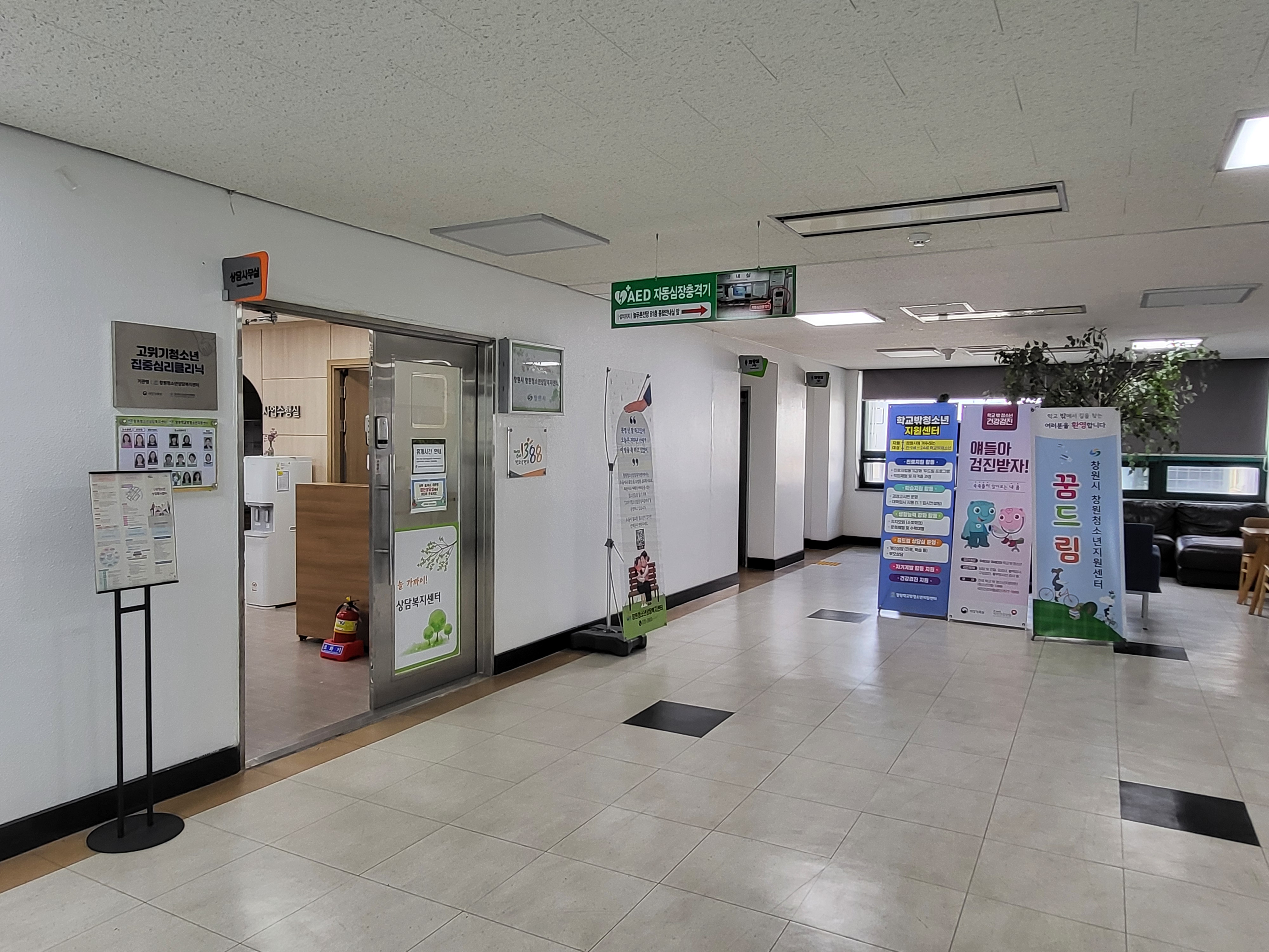 로비에서 바라본 창원시 청소년 상담 지원센터 입구를 촬영한 사진이며, 입구 좌측에 직원 및 프로그램 관련 안내판이 비치되어 있으며 우측에는 화장실과 휴식공간을 포함한 시설이 위치해 있습니다.  