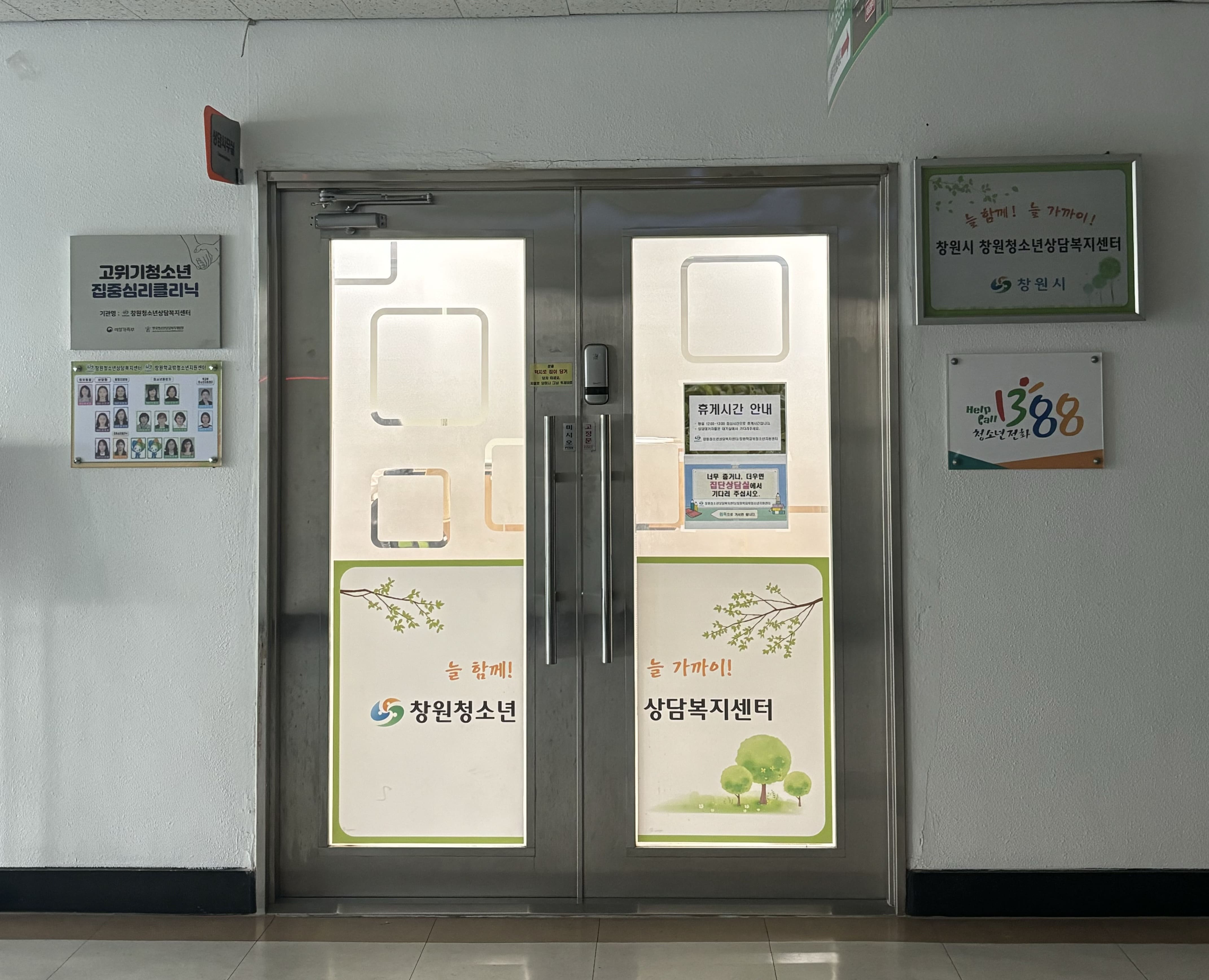 입구 앞 정면에서 바라본 창원시 청소년 상담복지센터 입구를 촬영한 사진입니다. 입구 좌측에는 직원 조직도 및 고위기 청소년 집중심리클리닉 팻말이, 우측에는 창원시 청소년 상담복지센터 팻말과 청소년전화 1388 팻말이 설치되어 있습니다.