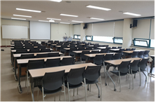 교육실2 뒤 가장자리에서 전체를 바라보며 촬영한 사진입니다. 2인용 책상 3열로 놓여있고 앞에 스크린이 내려와있습니다.