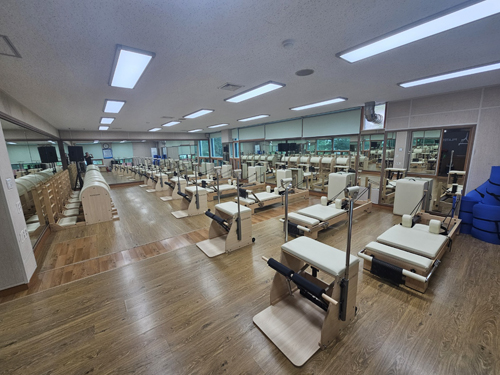 진해국민체육센터의 1층 기구 필라테스실 입구 쪽 가장자리에서 바라본 전체 사진이며, 좌측 유리 앞을 기준으로 바렐, 체어, 리포머 등 기구들이 일렬로 배치되어 있습니다.