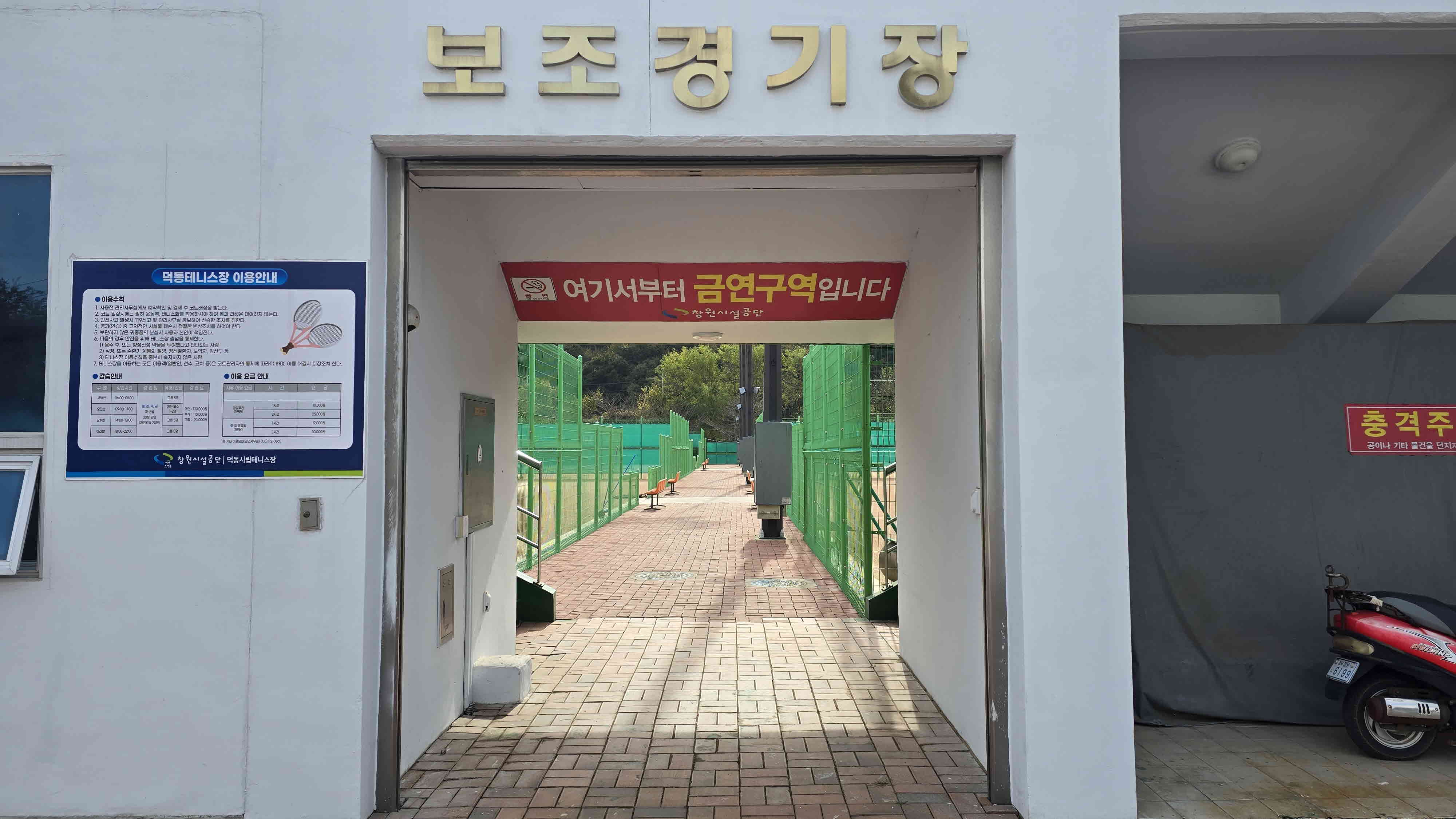 보조경기장으로 들어가는 입구를 담은 사진이며, 입구 좌측에 요금 안내 등 안내 표지판이 게시되어 있고 보조경기장 내에서는 금연구역이라는 안내 문구도 입구 위에 게시되어 있습니다.