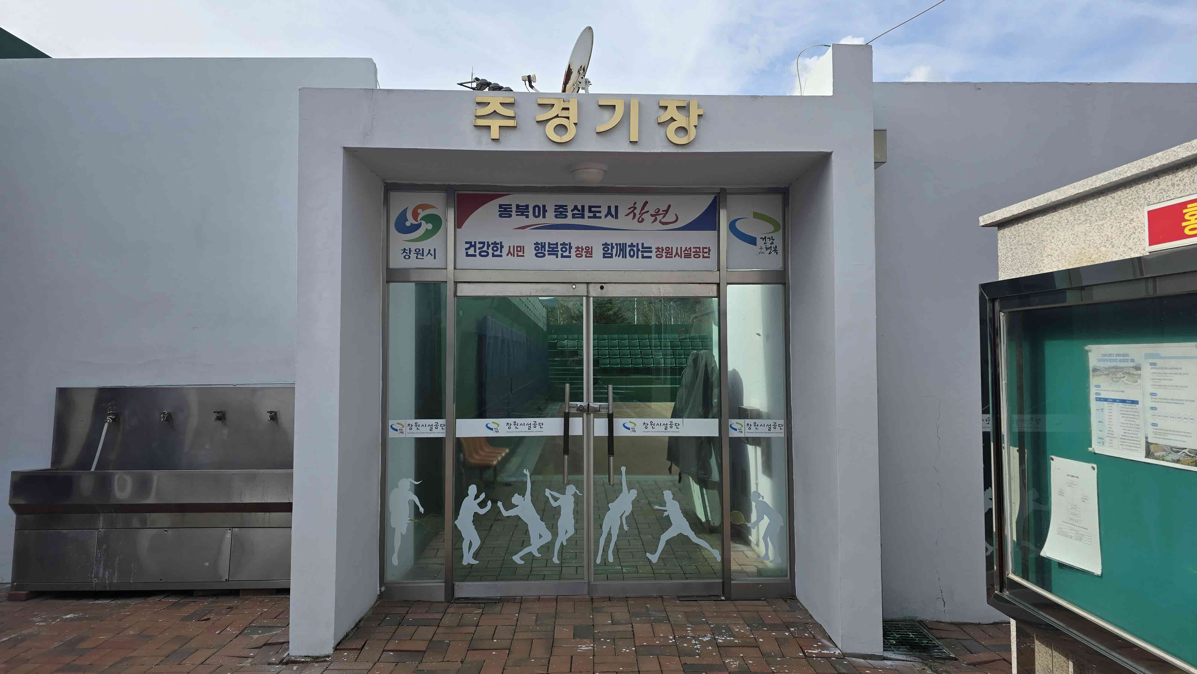 주경기장 입구를 담은 사진이며, 입구를 기준으로 좌측에는 음수대가 있고 우측에는 게시판이 비치되어 있습니다.